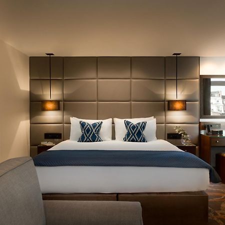 Montcalm Chilworth Townhouse, Paddington Hotell London Eksteriør bilde The photo shows a modern hotel room featuring a neatly made bed with two decorative pillows at the headboard. The headboard itself is upholstered in a grid pattern and flanked by wall-mounted lamps. There's a small desk area to the side with a mirror