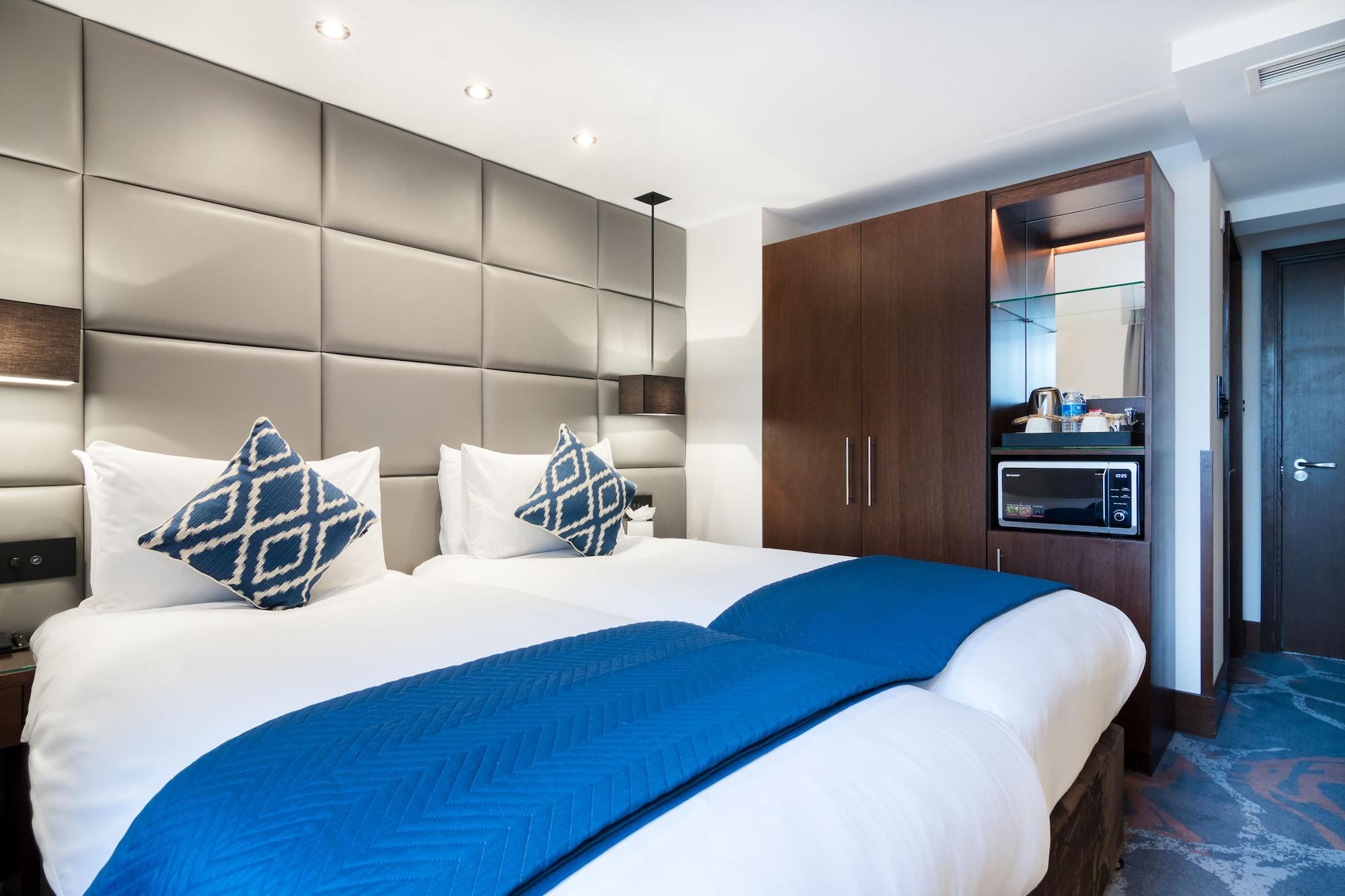 Montcalm Chilworth Townhouse, Paddington Hotell London Eksteriør bilde The photo shows a modern hotel room featuring two beds with white linens and decorative blue throw blankets. Each bed has a pair of patterned cushions. The walls behind the beds are adorned with a sleek, padded design. To the side, there’s a wooden c