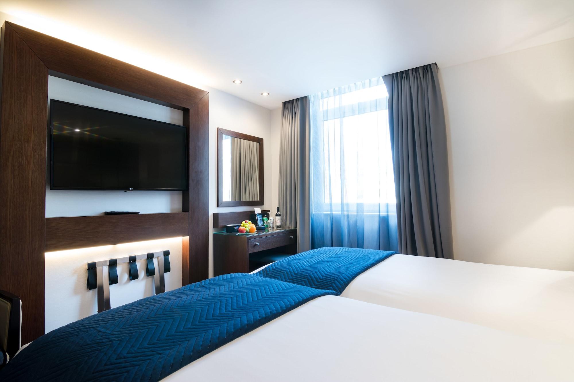 Montcalm Chilworth Townhouse, Paddington Hotell London Eksteriør bilde The photo shows a modern hotel room featuring two twin beds with white linens and deep blue throws. There is a flat-screen television mounted on the wall opposite the beds, along with a mirror and a small dresser or desk. The room has large windows d