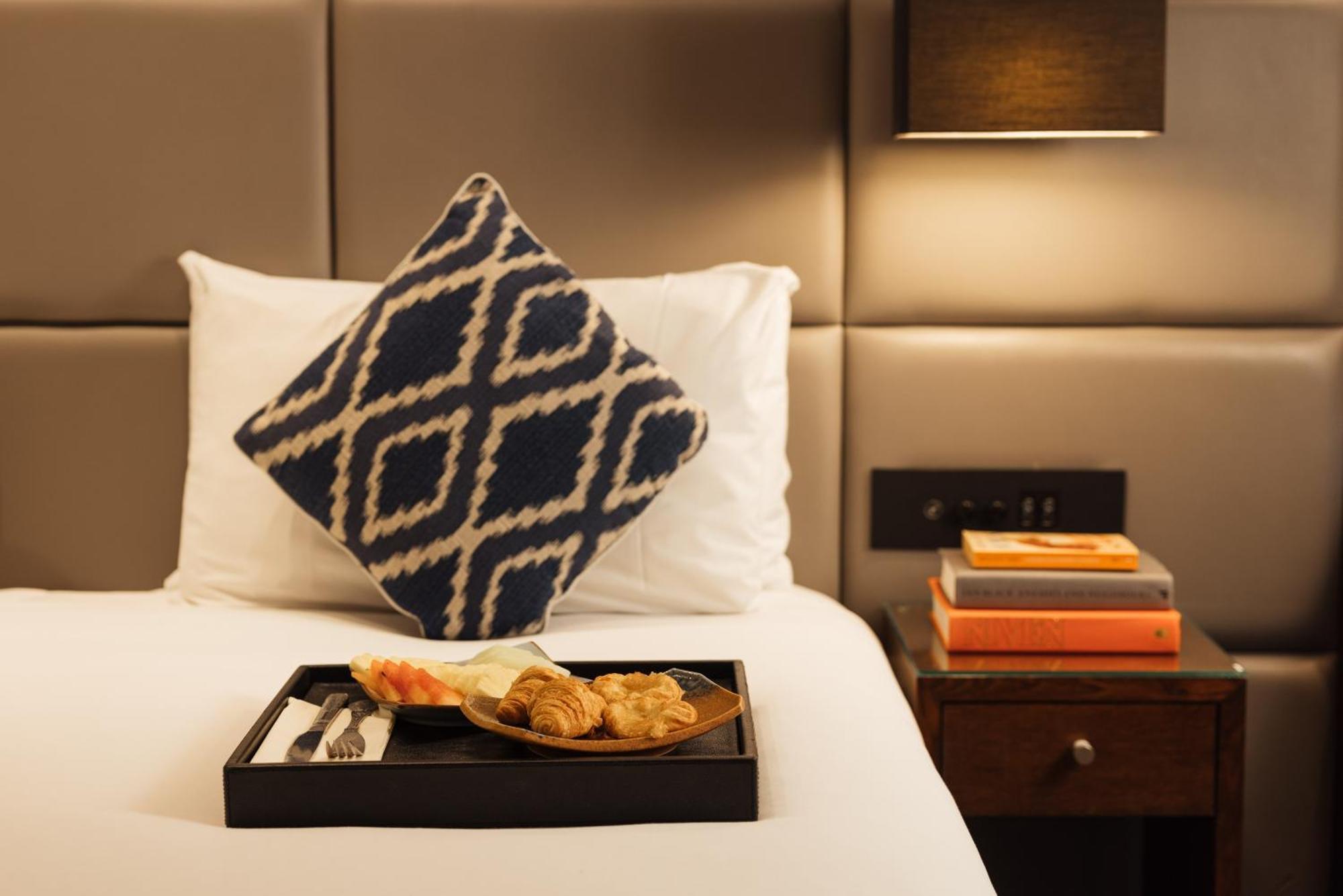 Montcalm Chilworth Townhouse, Paddington Hotell London Eksteriør bilde The photo shows a neatly arranged bed with a decorative pillow featuring a blue and white geometric pattern. On a tray placed on the bed, there are various food items, including pastries. Beside the bed, there is a small wooden nightstand with a few 