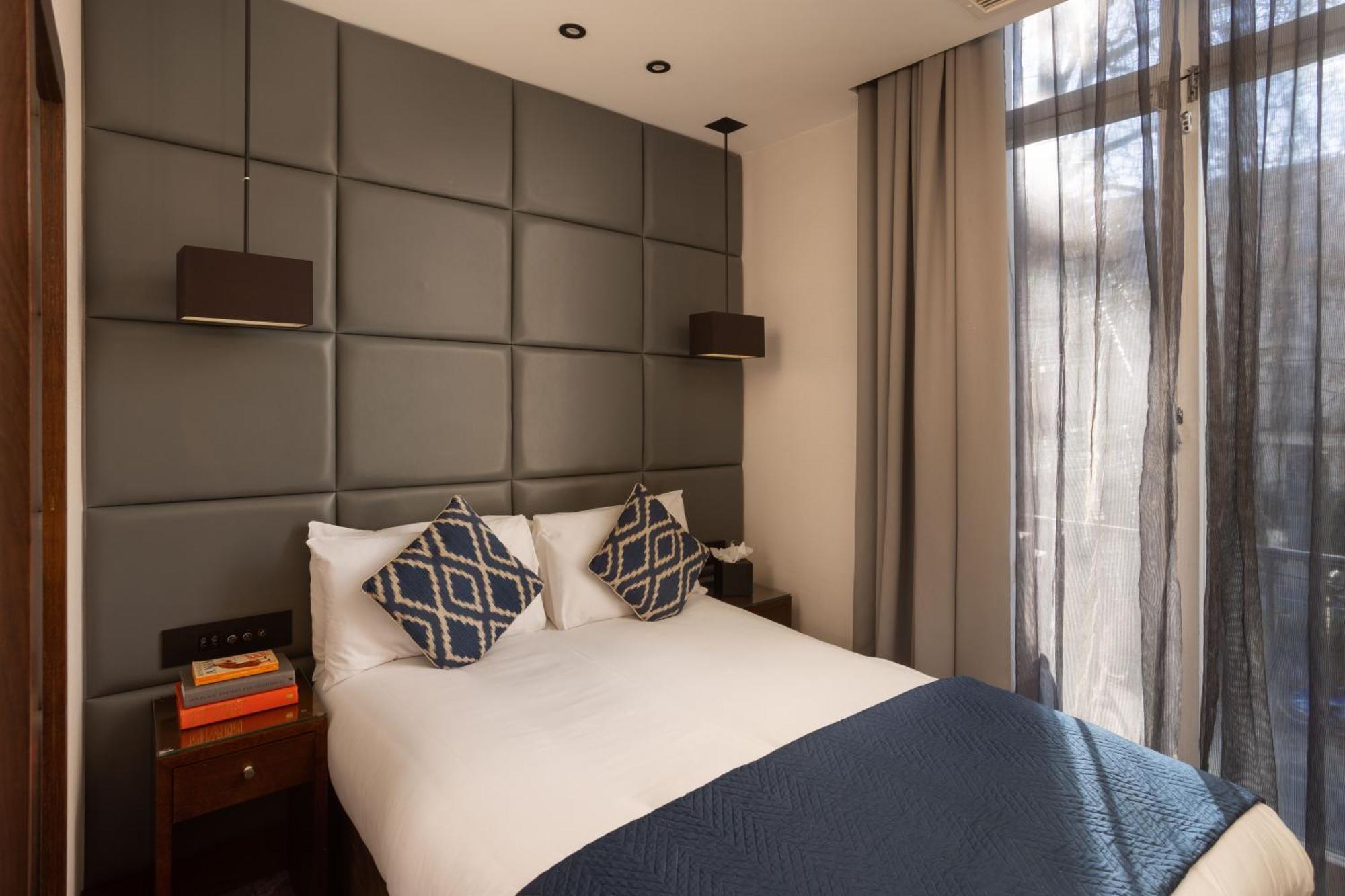 Montcalm Chilworth Townhouse, Paddington Hotell London Eksteriør bilde The photo shows a modern and cozy bedroom. The room features a double bed with white bedding and decorative pillows with a geometric pattern. Behind the bed is a stylish upholstered wall in a muted tone. There are two bedside tables, each with a lamp