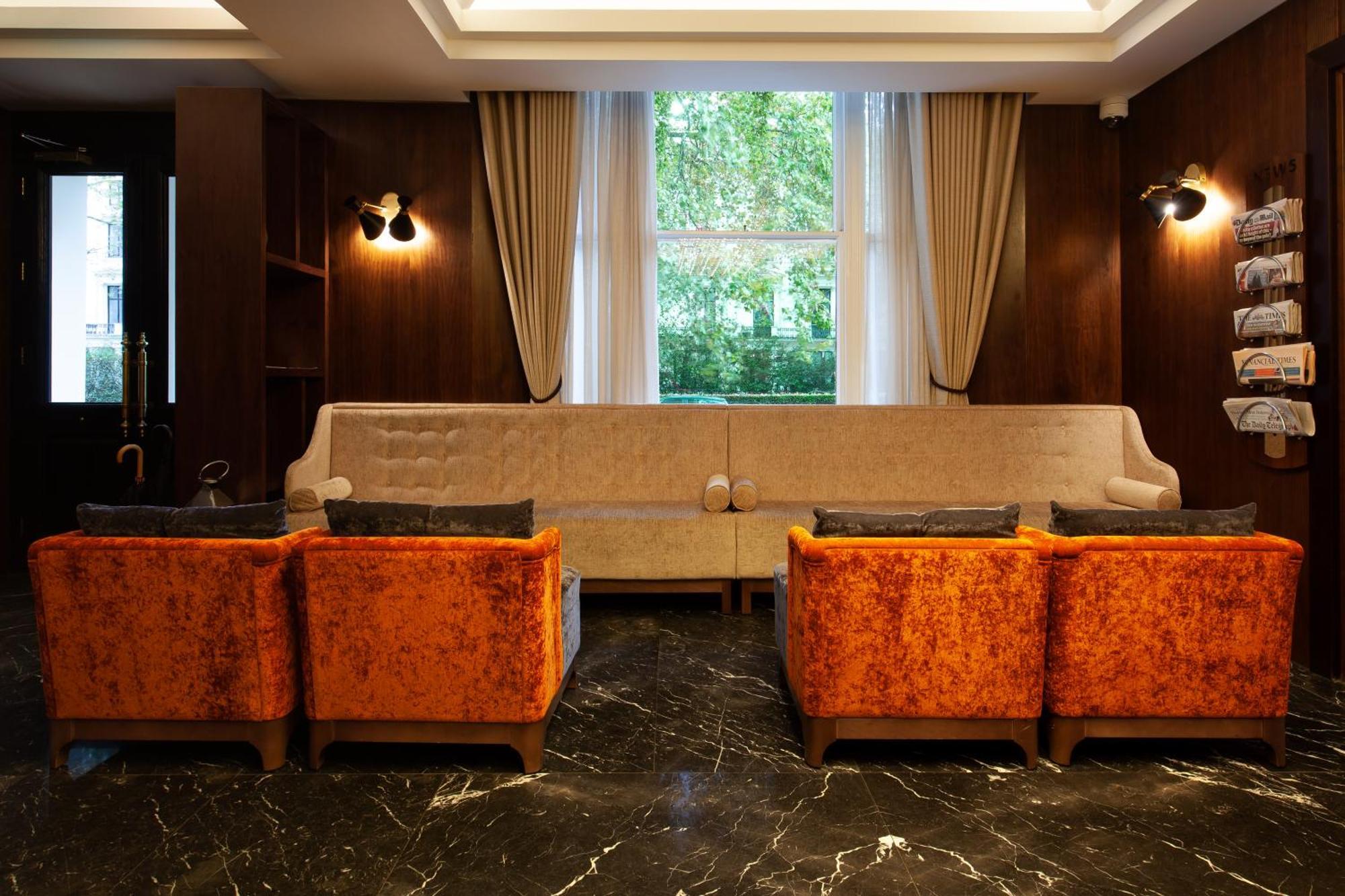 Montcalm Chilworth Townhouse, Paddington Hotell London Eksteriør bilde The photo shows a stylish interior space, likely a lounge or sitting area. It features a long, light-colored sofa against a wall with large windows that allow natural light to enter. On either side of the sofa, there are two orange, plush armchairs. 