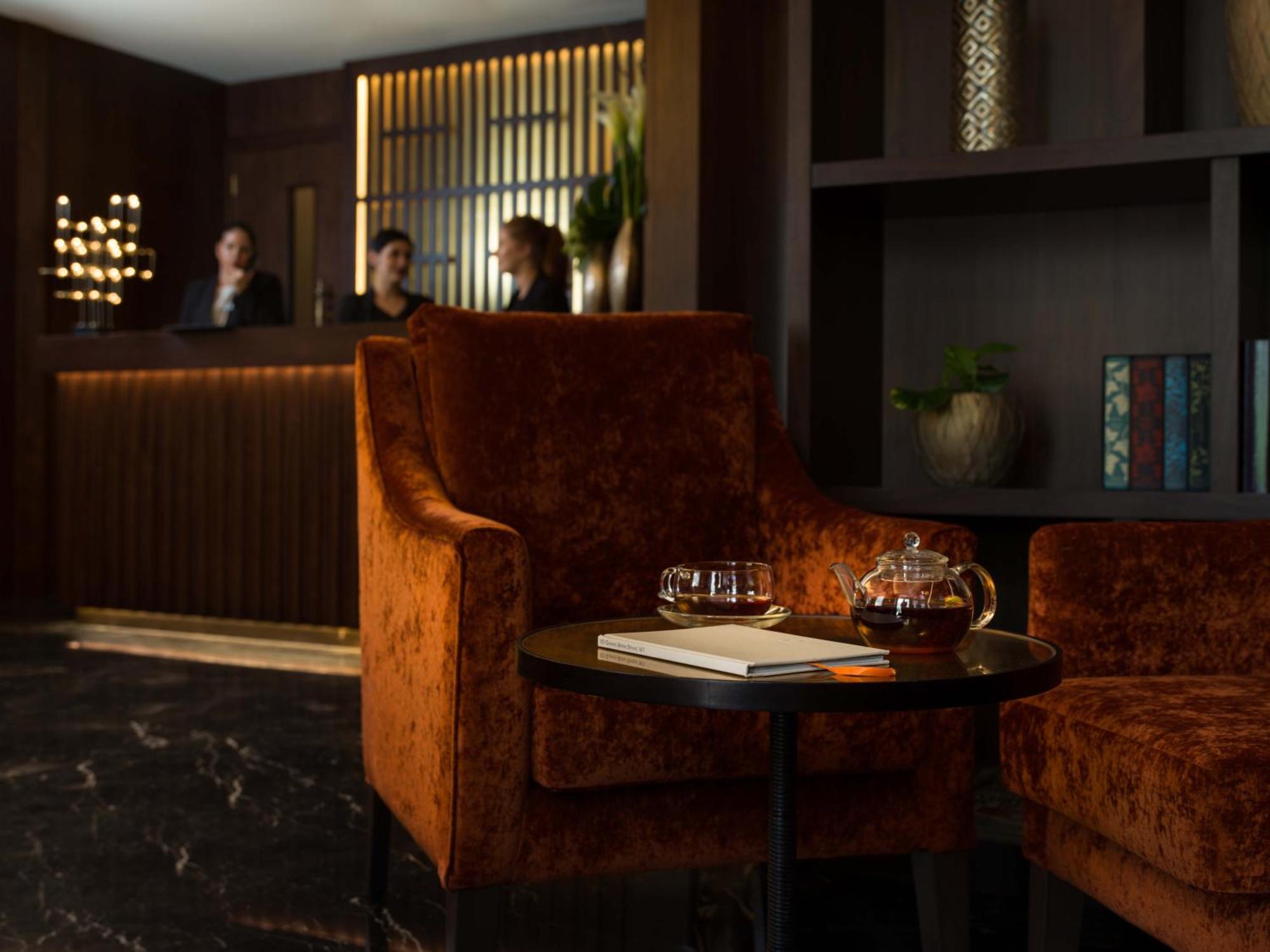 Montcalm Chilworth Townhouse, Paddington Hotell London Eksteriør bilde The photo depicts a cozy interior, likely part of a hotel lobby or lounge area. In the foreground, there are two plush orange chairs facing a small round table. On the table, there's a teapot, glasses, and a book, suggesting a relaxed atmosphere cond