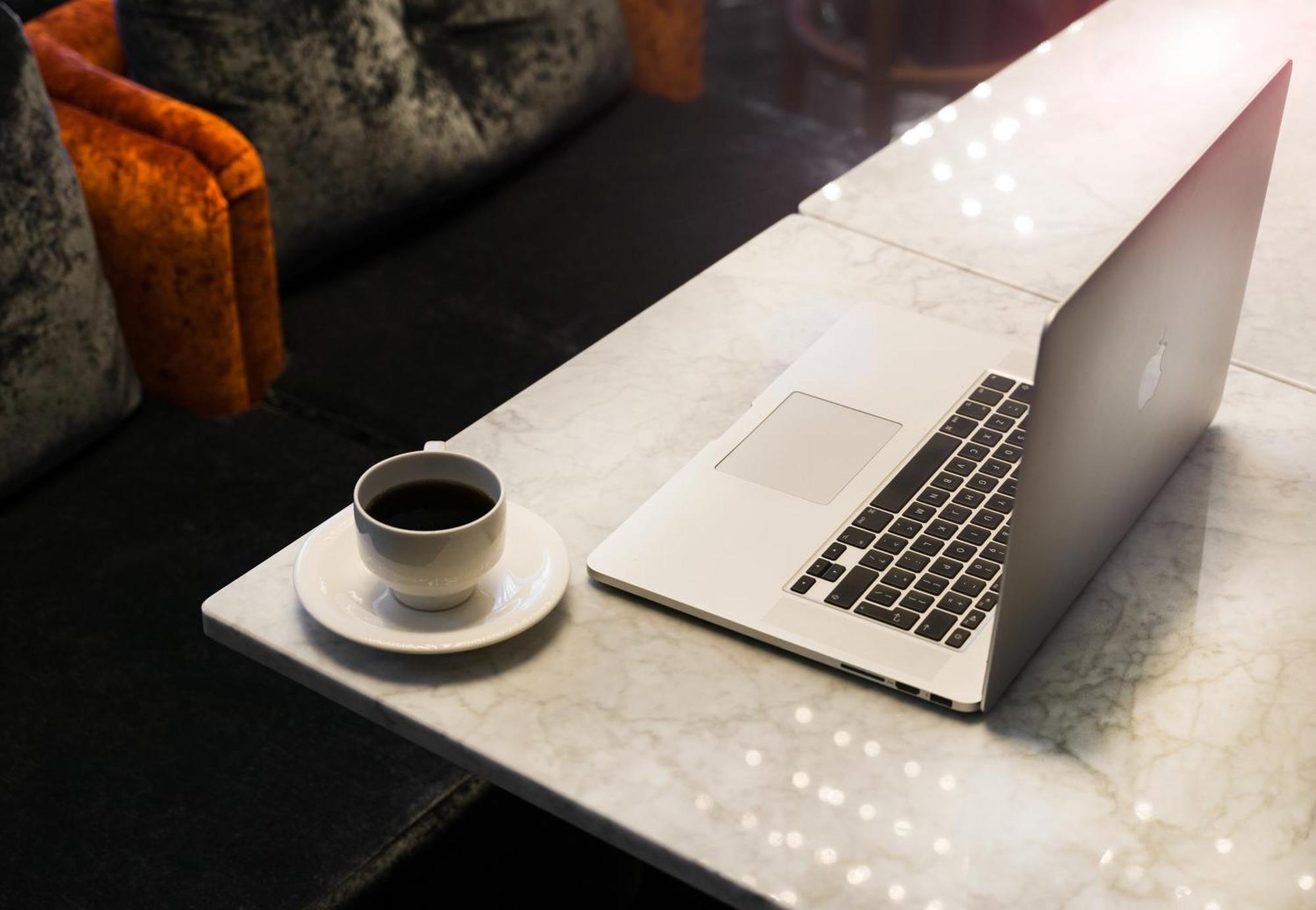 Montcalm Chilworth Townhouse, Paddington Hotell London Eksteriør bilde The photo shows a marble table with a laptop positioned on one side. Next to the laptop, there is a small cup of black coffee placed on a saucer. The background features a cozy seating area, with some upholstered chairs that have vibrant orange and d