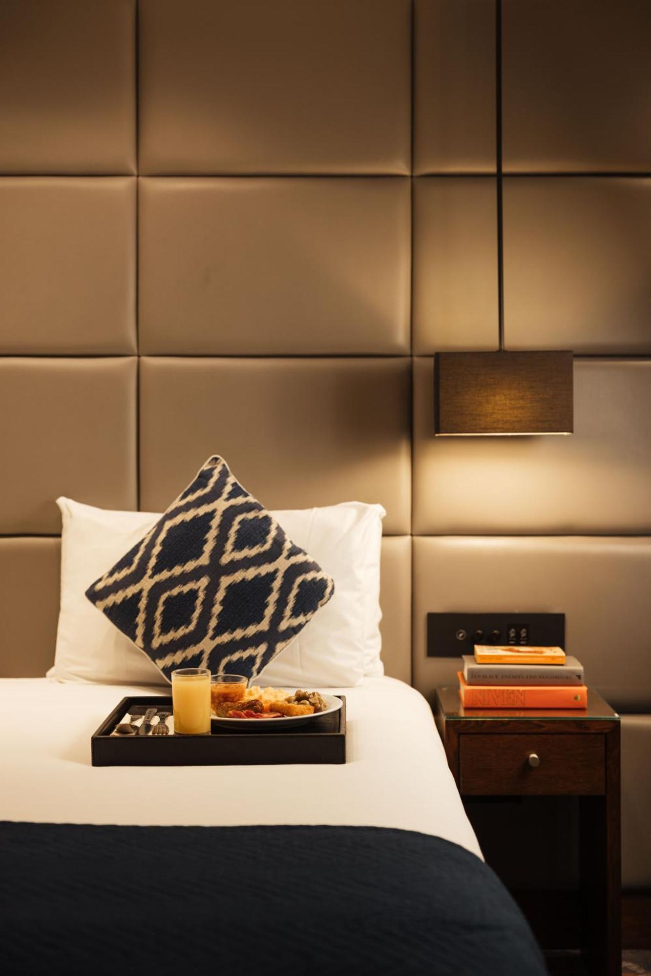 Montcalm Chilworth Townhouse, Paddington Hotell London Eksteriør bilde The photo shows a cozy hotel room setting with a neatly made bed featuring white bedding. A decorative pillow with a blue and white geometric pattern is propped on the bed. There is a bedside table with a black lamp, and next to it, there are some bo