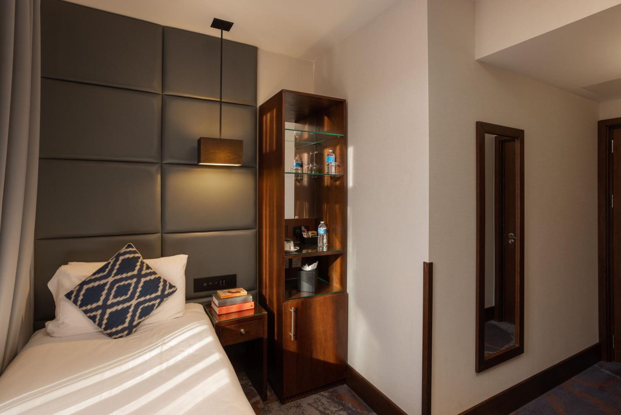 Montcalm Chilworth Townhouse, Paddington Hotell London Eksteriør bilde The photo shows a small, modern hotel room. On the left side, there is a single bed with a decorative pillow featuring a blue and white geometric pattern. Above the bed is a dark panel with a lamp attached. 

Next to the bed, there is a wooden nights
