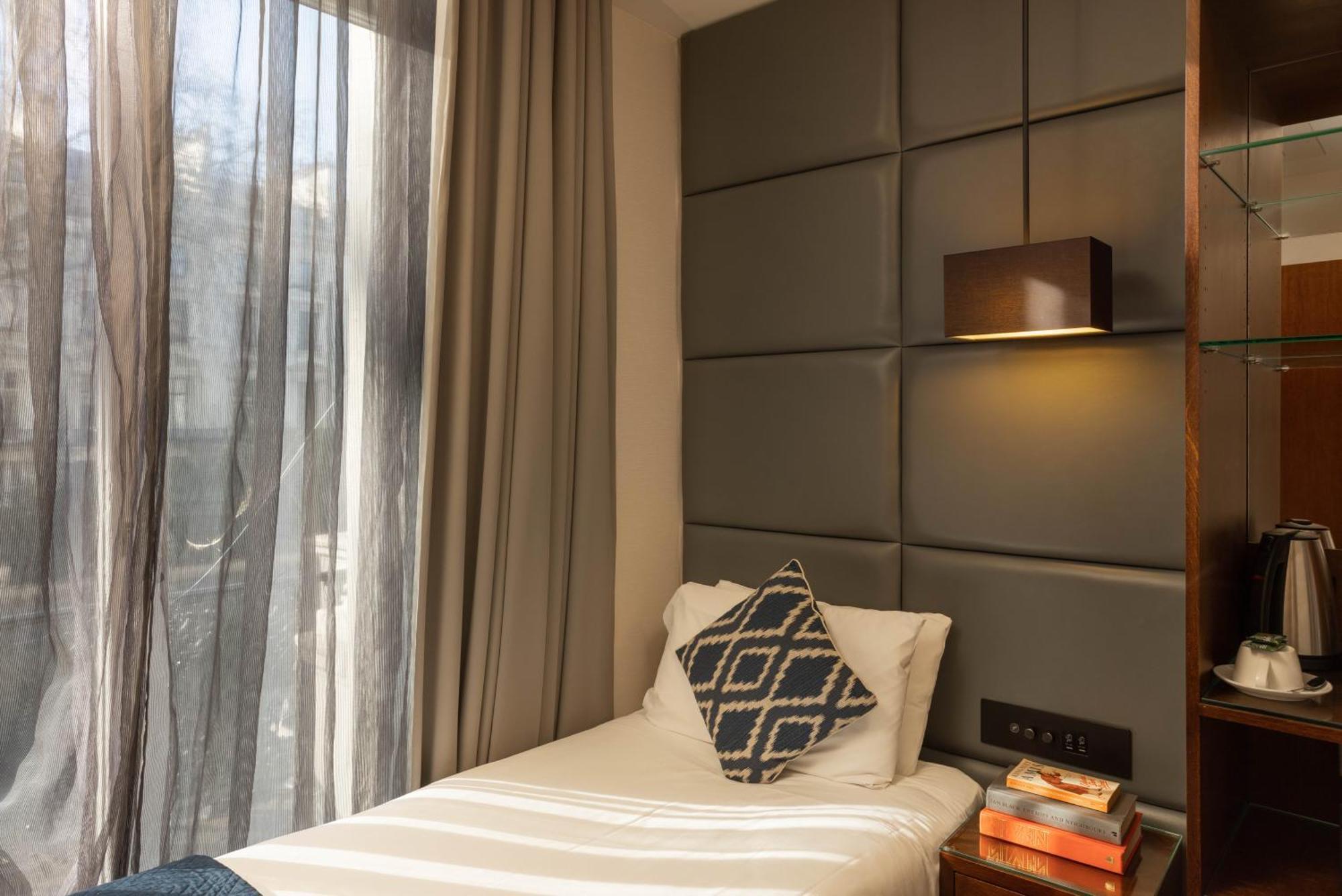 Montcalm Chilworth Townhouse, Paddington Hotell London Eksteriør bilde The photo shows a small, modern bedroom featuring a single bed with white bedding and a decorative pillow that has a geometric pattern. The wall behind the bed has a padded design, and there is a lamp mounted above the bed. Large curtains frame a win