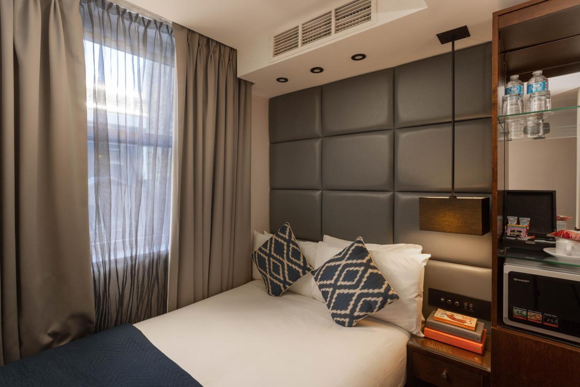 Montcalm Chilworth Townhouse, Paddington Hotell London Eksteriør bilde The photo shows a compact hotel room with a cozy design. The bed is made up with white linens and two decorative pillows featuring a geometric pattern. Behind the bed, there is a padded headboard in a neutral tone. Natural light filters through sheer