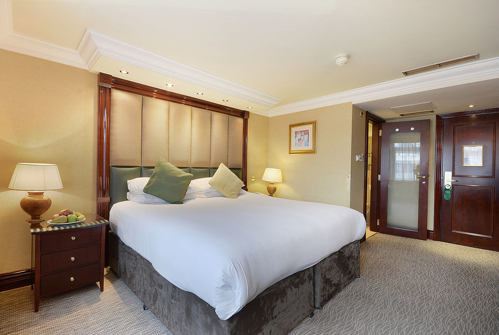 Montcalm Chilworth Townhouse, Paddington Hotell London Eksteriør bilde The photo shows a well-furnished hotel room. In the center is a large bed with a white duvet and two decorative green pillows. Beside the bed, there is a wooden nightstand with a lamp and a small bowl of fruits. The room features warm lighting and ne